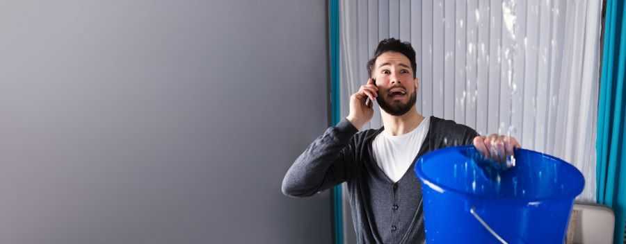 Man on the Phone With a Panicked Expression Holding a Blue Bucket Collecting Dripping Water