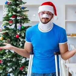 Man Standing in Front of Christmas Tree While Shrugging and Leaning on Crutches With a Black Eye Wearing a Neck Brace and a Santa Hat